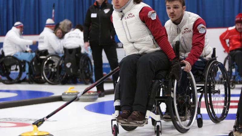 a female wheelchair curler from Latvia plays a stone while another curler holds her wheelchair to steady it