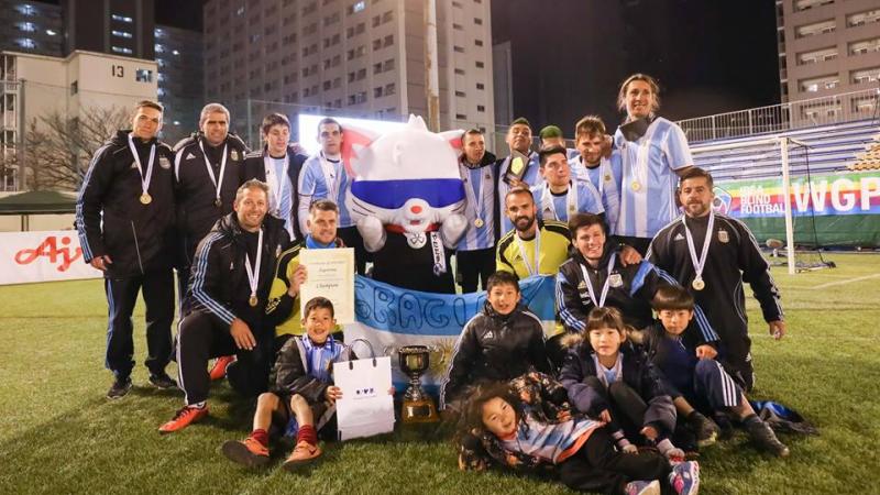 A group of male blind footballers stand with a blindfolded mascot holding a trophy and the Argentinian flag