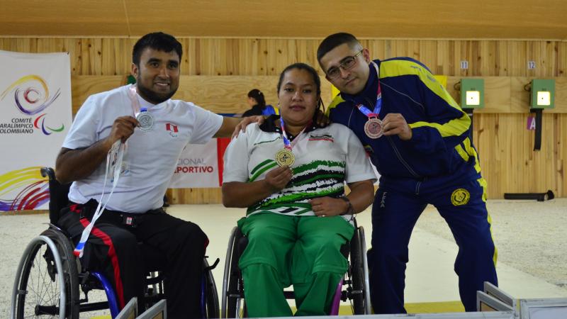 female Para shooter Maria Teresa Restrepo sitting a wheelchair with a male athlete either side holding up medals