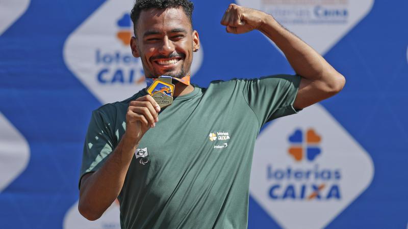 male Para athlete Daniel Martins holds up his arm and fist pumps while holding a gold medal and smiling