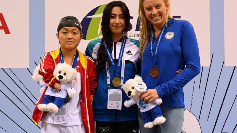 three female swimmers on the podium holding their medals and mascot teddys