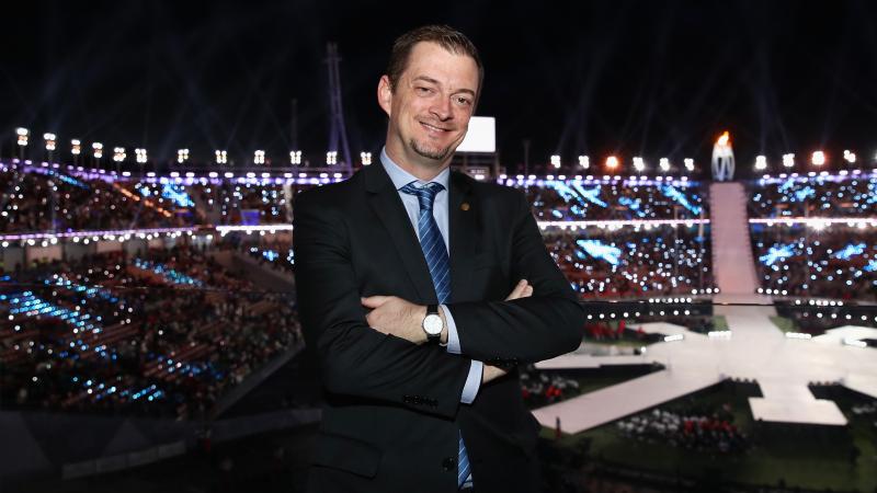 IPC President Andrew Parsons smiles and crosses arms with the PyeongChang 2018 stadium in the background during the Closing Ceremony