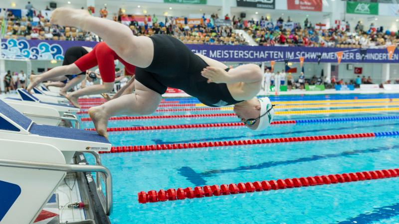 A short stature female swimmer jumping in the water in front of other swimmers