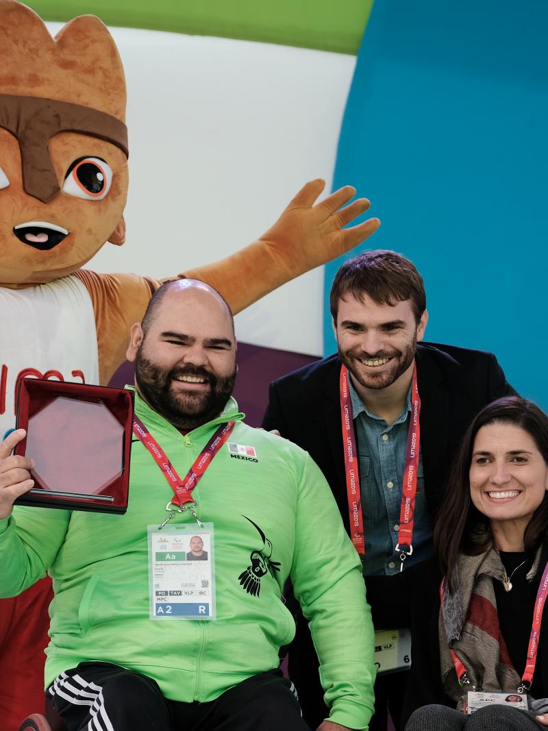 A male powerlifter holds up an award smiling with two other people and a furry mascot standing behind