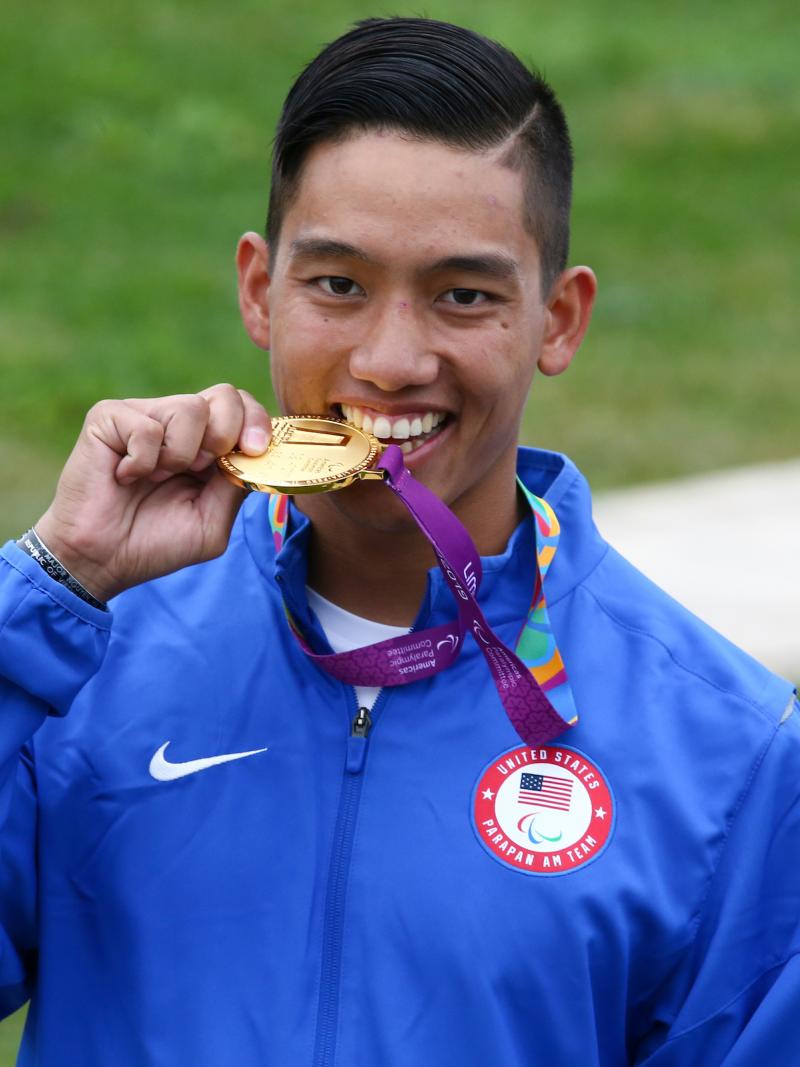 a male Para shooter biting a gold medal and smiling
