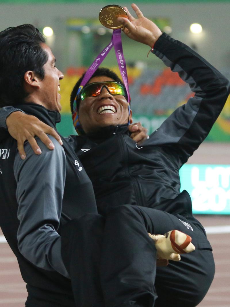 a male vision impaired runner being carried by his guide while waving his medal