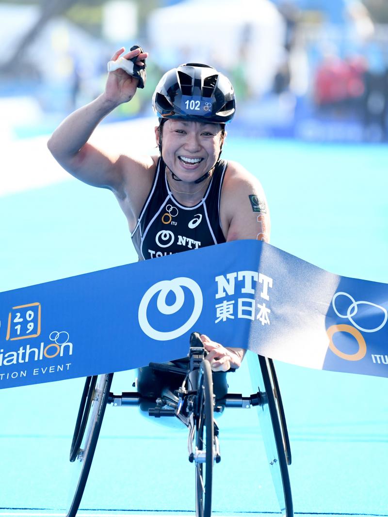 a female Para triathlete in a wheelchair raises her arm as she breaks the finish line tape