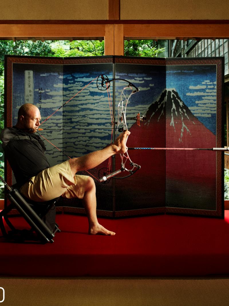 Armless archer sits down holding bow and arrow with foot with Japanese backdrop