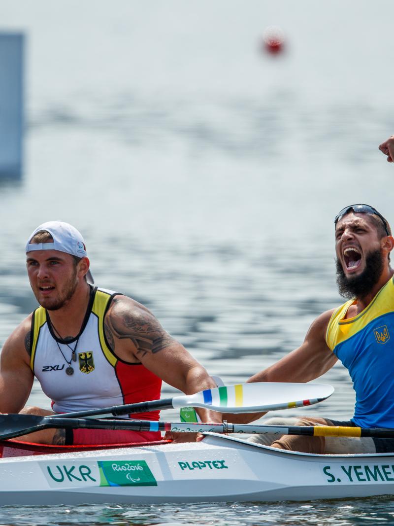 two male Para canoeists side by side in their boats with one punching the air in celebration