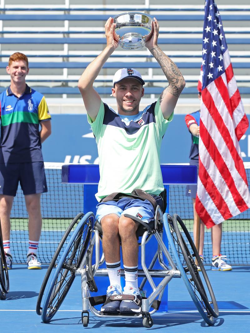 a male wheelchair tennis player holds up a trophy