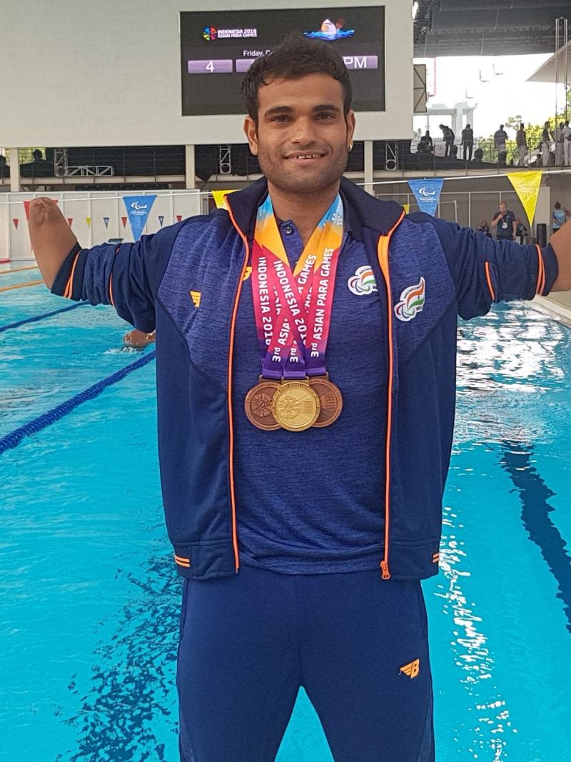 a male Para swimmer standing in front of a swimming pool with medals round his neck