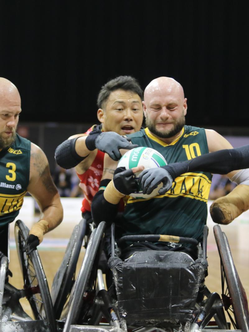 Man tries to hold onto wheelchair rugby ball while defenders surround him