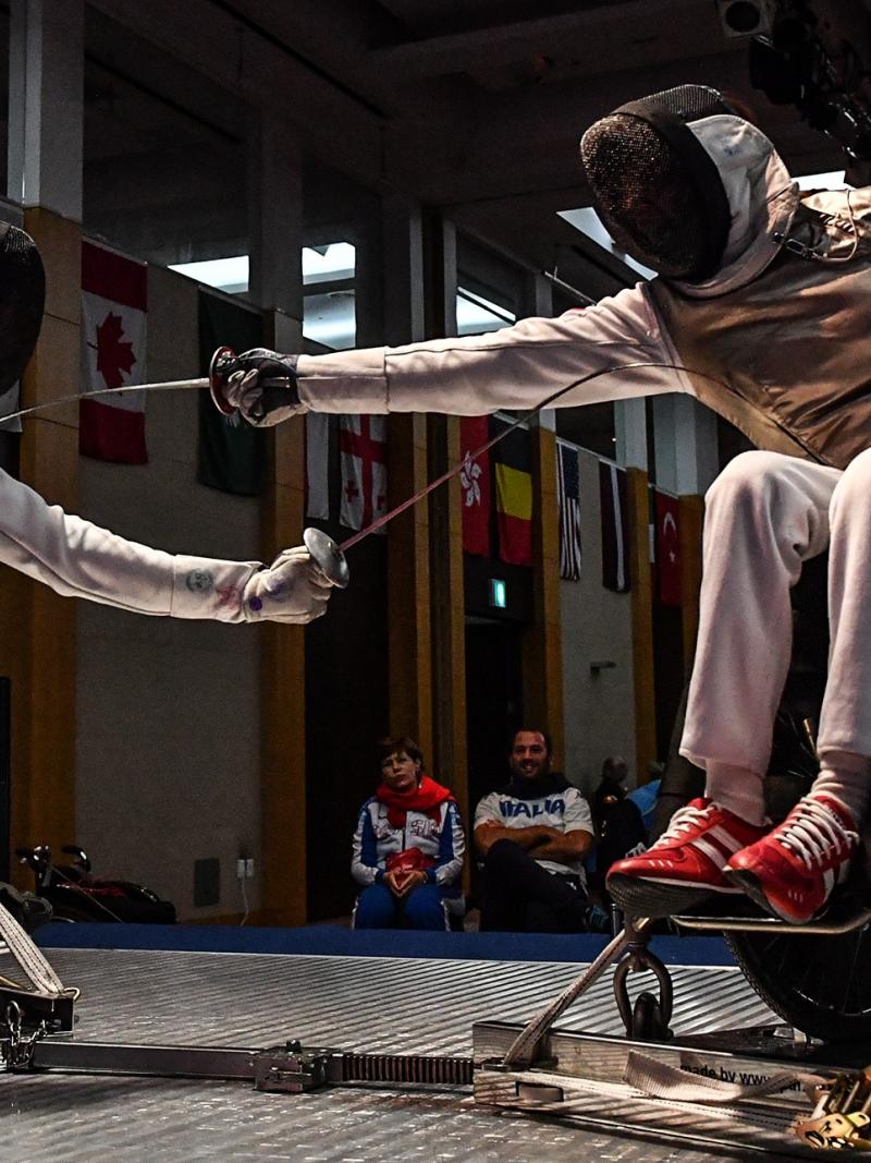 Two wheelchair fencers reach to strike their opponent