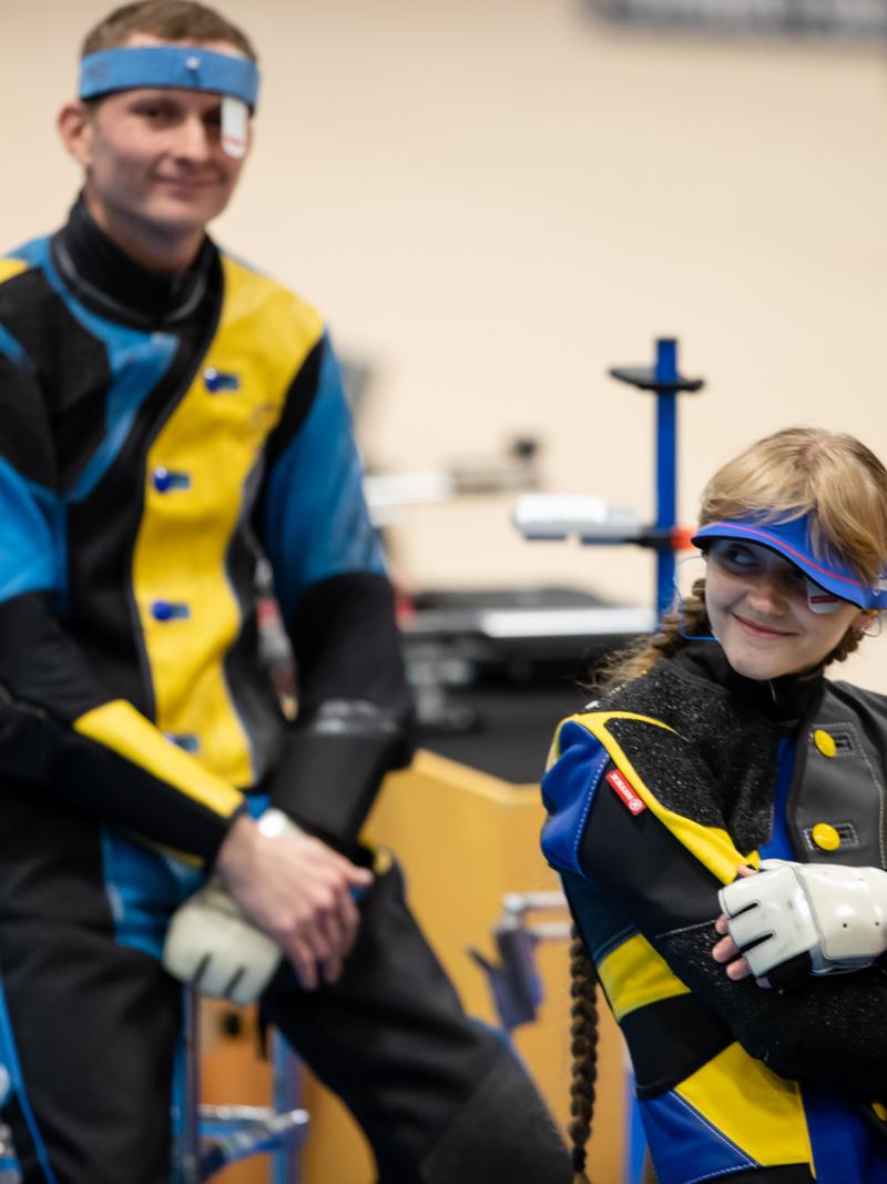 Ukrainian female shooter sits down smiling with male teammate behind also smiling