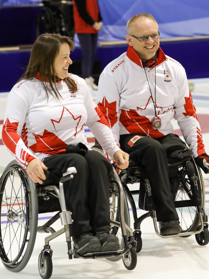 Man and woman in wheelchairs smile on curling ice sheet