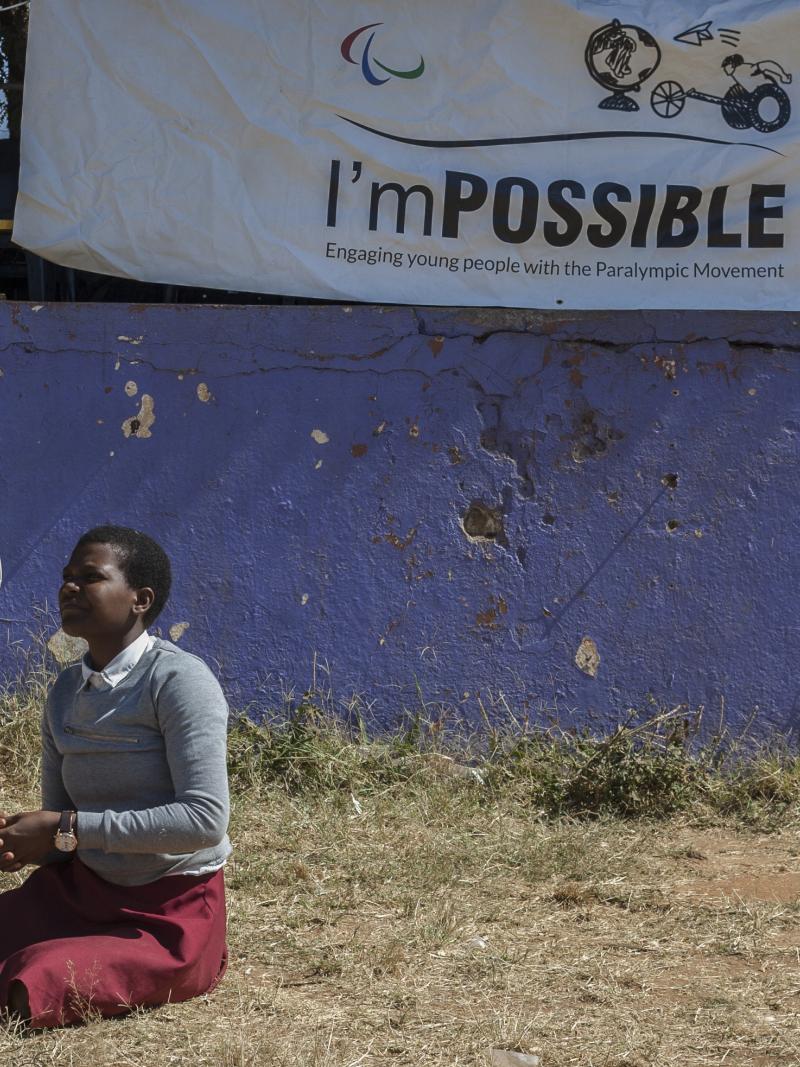 Student sits on the grass during an I'mPOSSIBLE activity in Malawi, with a flag of the programme in the background