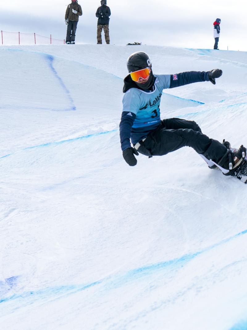 A male Para snowboarder competing being observed by eight people in the background