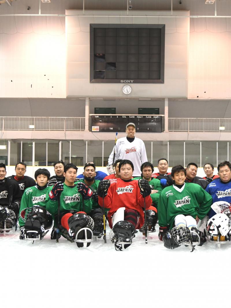Two men standing on a ice rink behind a group of 20 Para ice hockey players on sledges