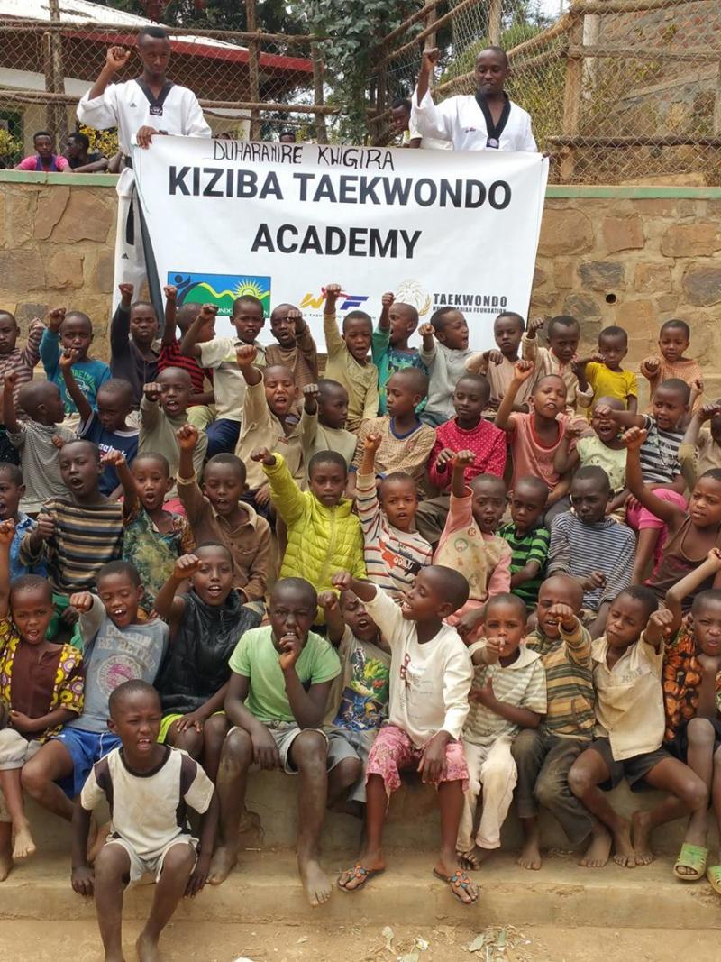 Group of African kids posing with fists raised and a banner behind saying Kiziba Taekwondo Academy