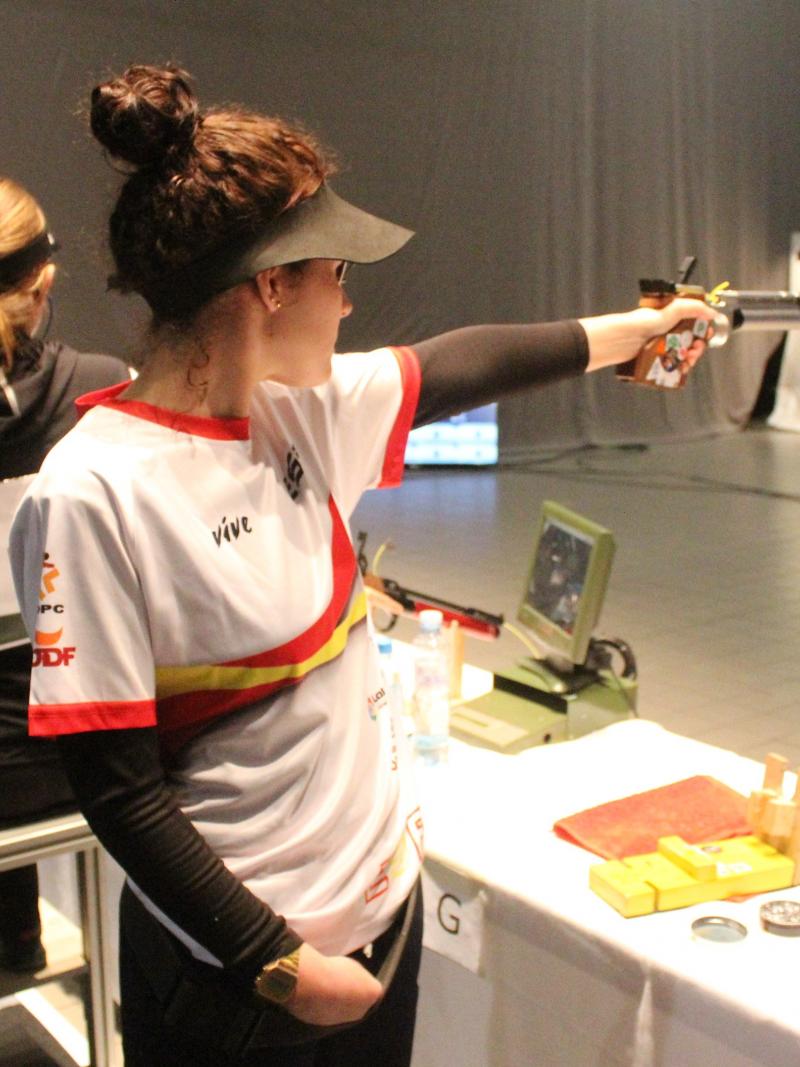 A woman standing and pointing a pistol to a target in an indoor shooting range with four women in the background