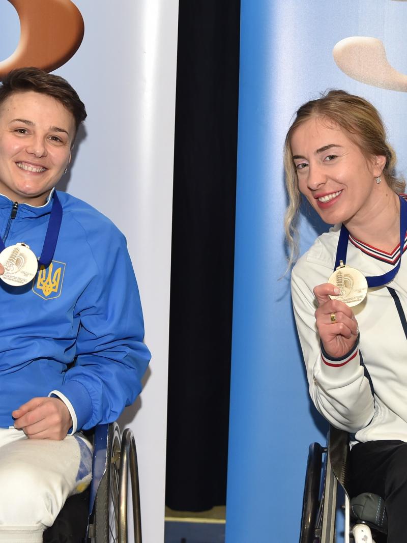 Two female wheelchair fencers pose with their medals on the podium