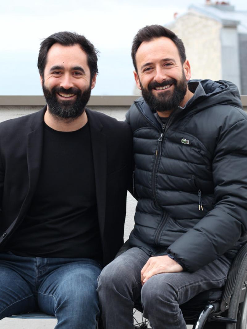 Two French males, one on a wheelchair, take a photo together smiling
