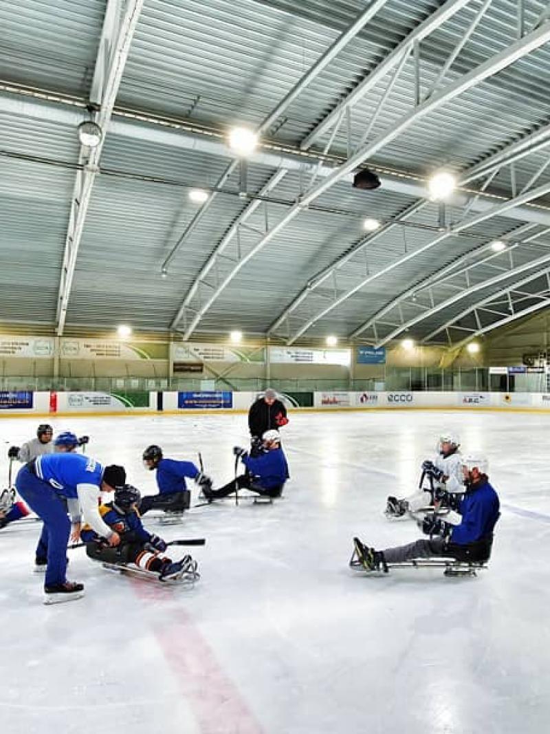 A group of eight persons on sledge hockey receiving instructions from three standing persons on an ice rink