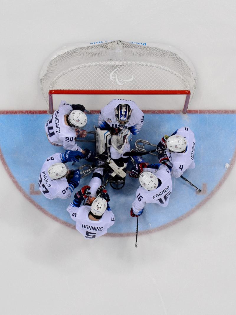 A group of six Para ice hockey players huddle together in front of the goal