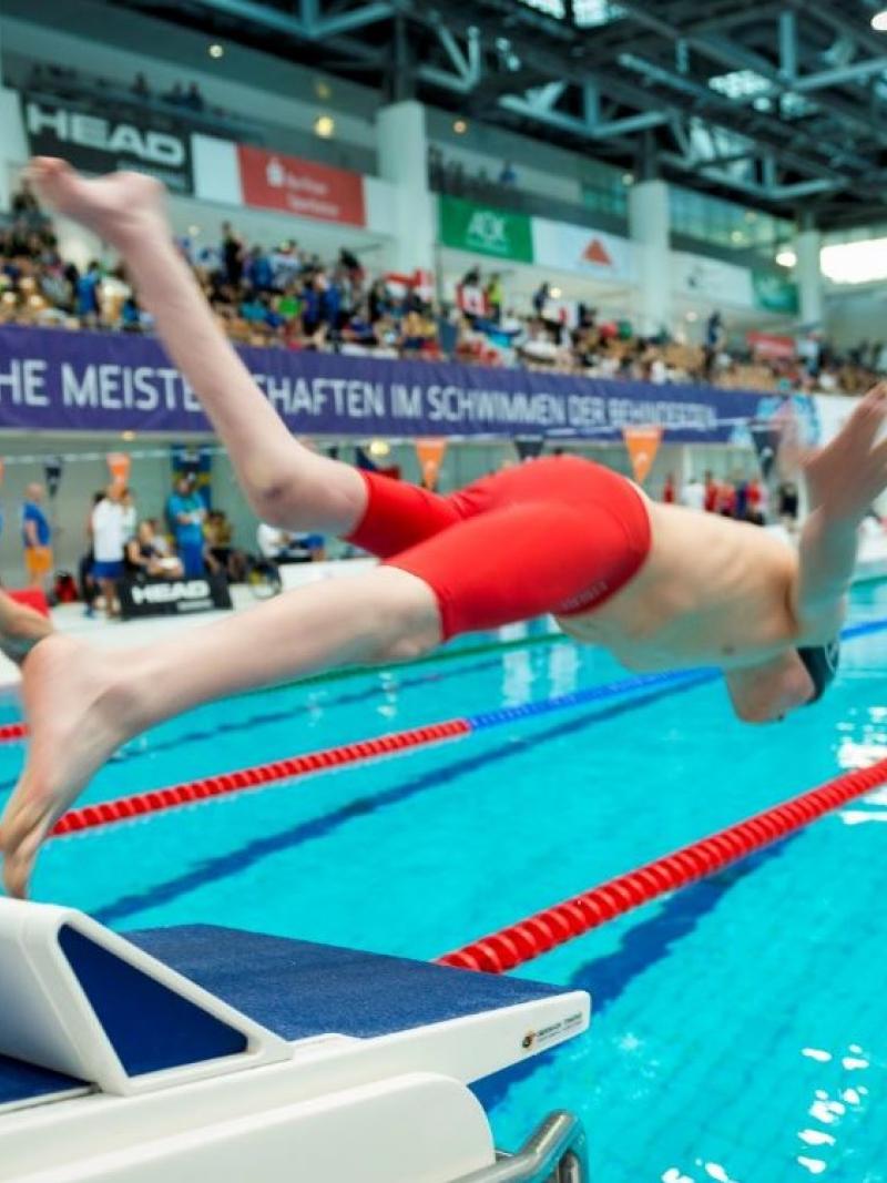 Two men jumping in the pool from starting blocks with people watching them from the stands