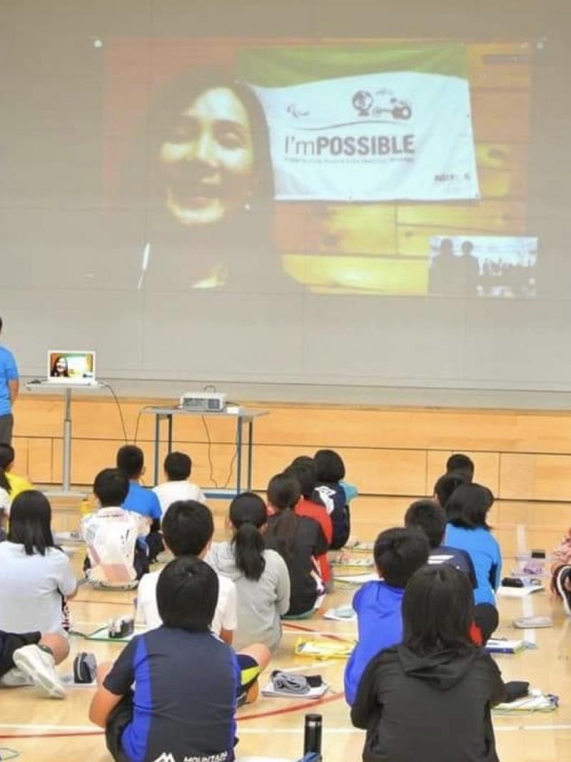 School children in a gym looking at a screen with a woman speaking via video call