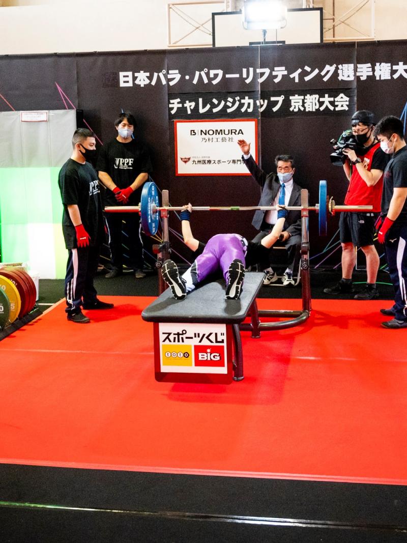 A person on a bench press during a Para powerlifting competition observed by three judges, three loaders and two cameramen