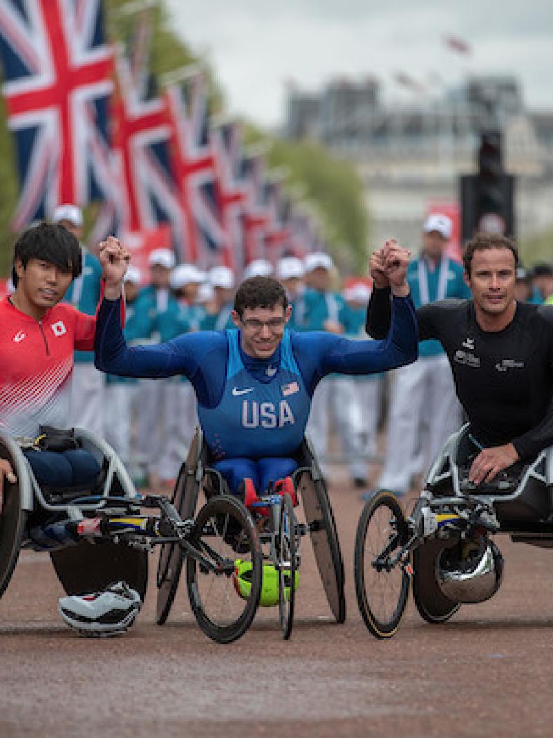 Three men in racing wheelchairs holding hands with the public and flags of the United Kingdom behind them