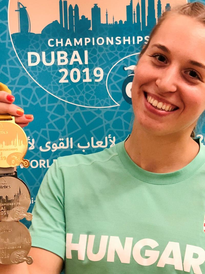 A young woman holding three medals and smiling with a shirt written Hungary