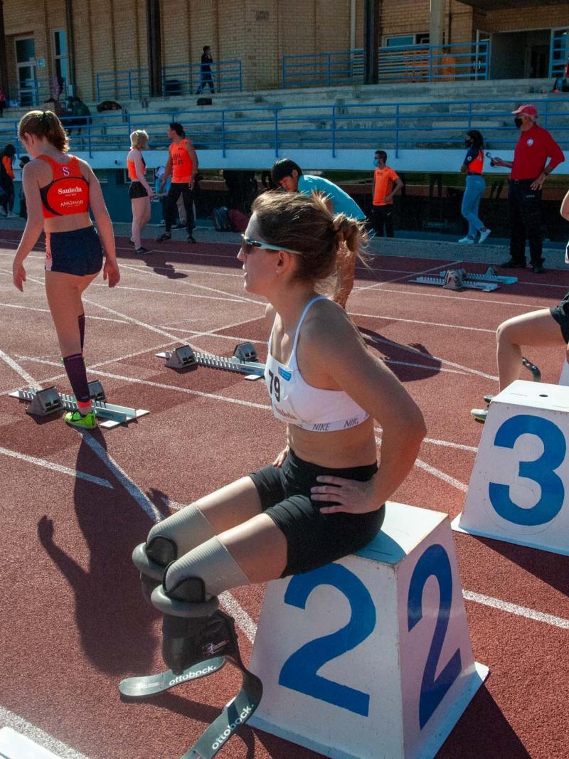 Two women with prosthetic legs sitting on starting blocks on a red athletics track with two other women standing in front of them