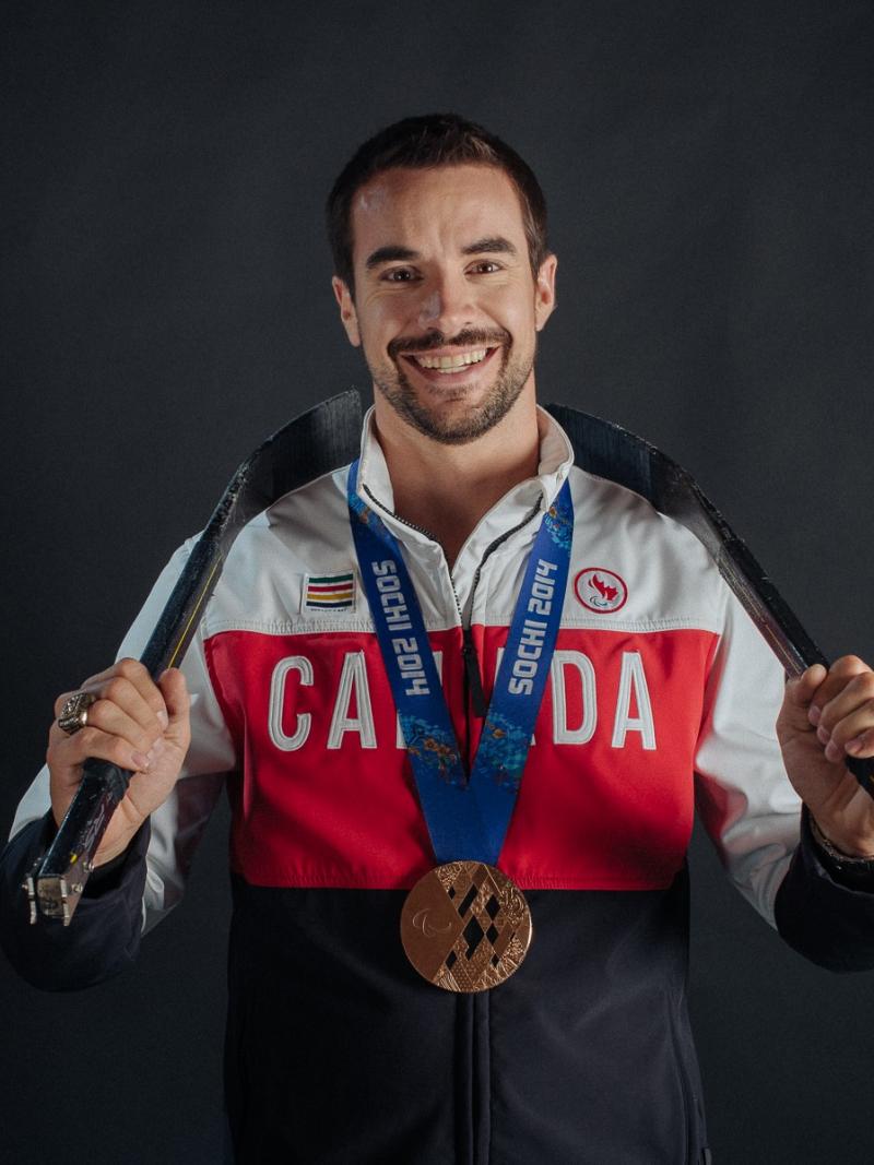 A smiling man with a bronze medal and wearing a Canadian jersey holds hockey sticks behind his neck