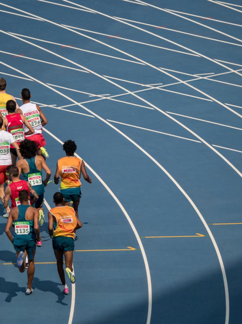 A group of 14 men seen from behind running in a blue athletics track