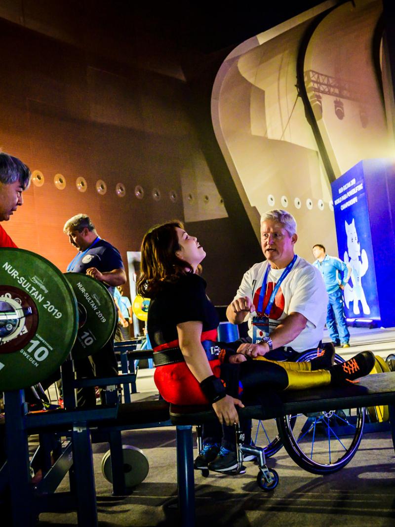 A woman on a bench press talking to a man in a wheelchair right next to her while another man stands near the barbell