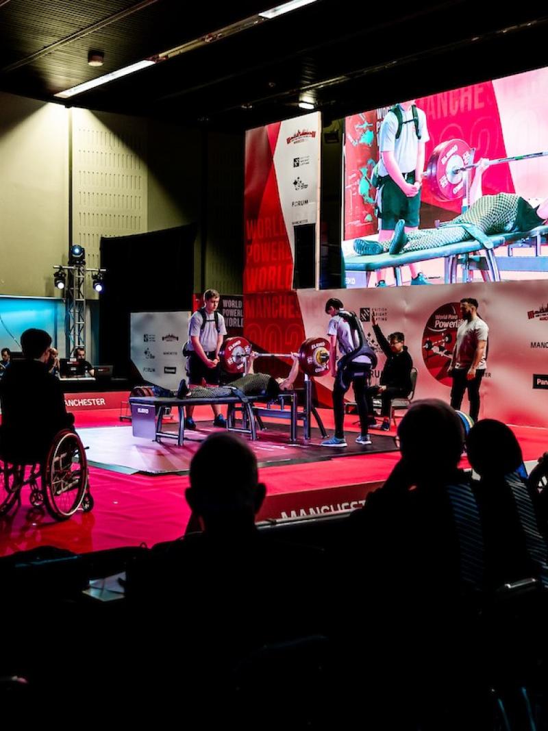 A group of people watching the stage of a Para powerlifting competition with a person competing surrounded by five people