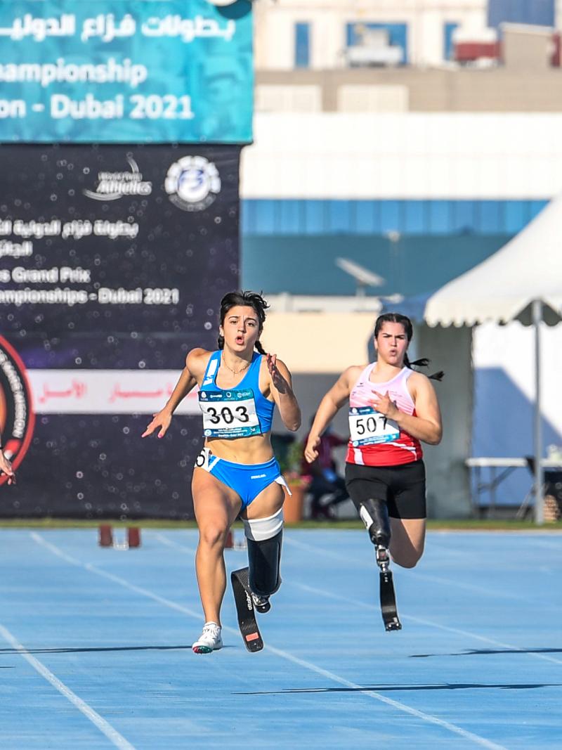 A group of five women with prosthetic legs running on a blue athletics track