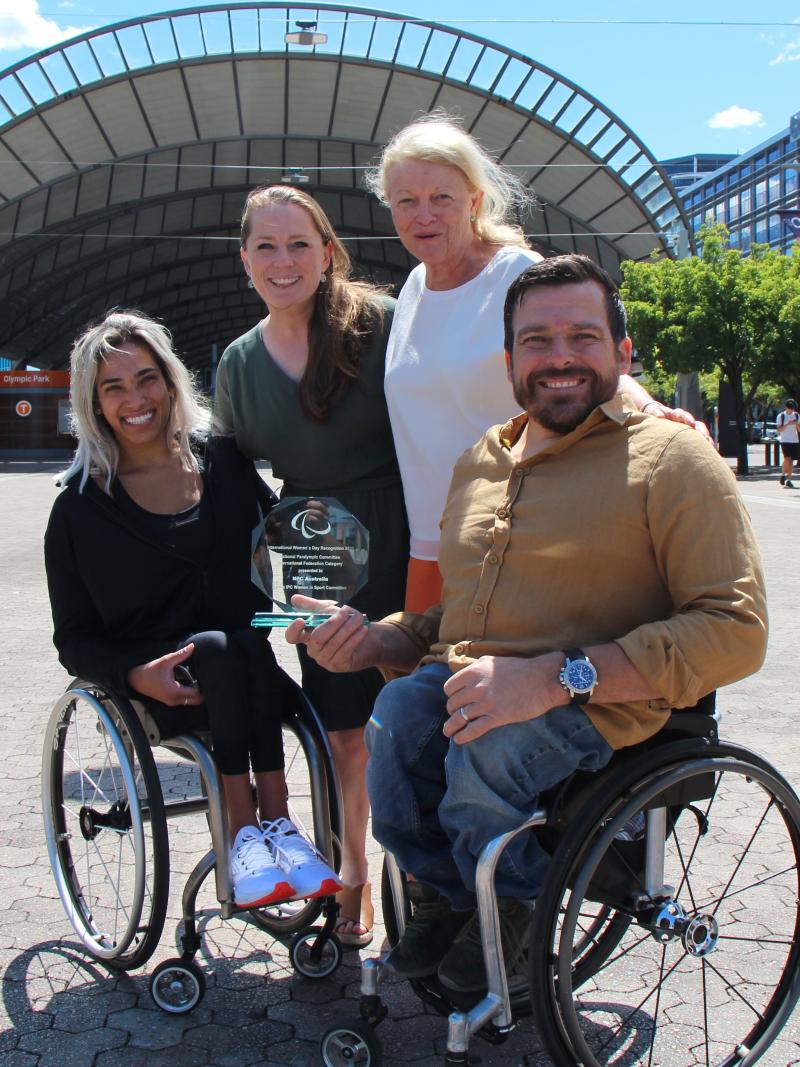 Two people in a wheelchair and two women standing smile with an award