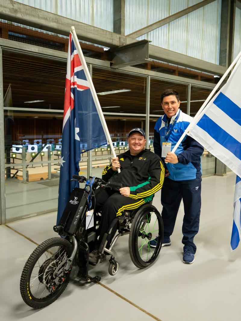 A man in a wheelchair holding the Australian flag posing next to a man standing and holding the flag of Uruguay
