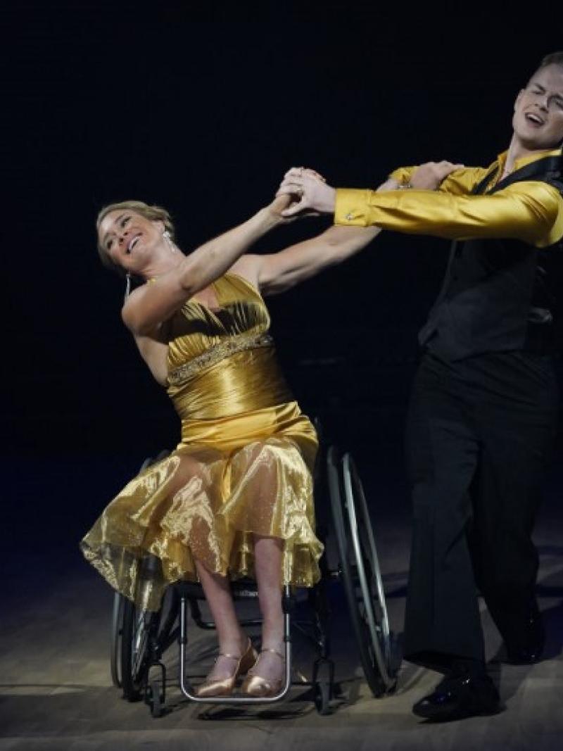 A female wheelchair dancer holding hands with her male standing dance partner on a dancefloor