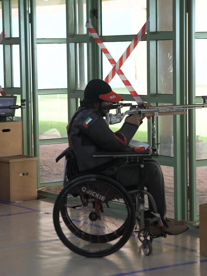 A man standing behind a man in a wheelchair shooting with a rifle in a shooting range