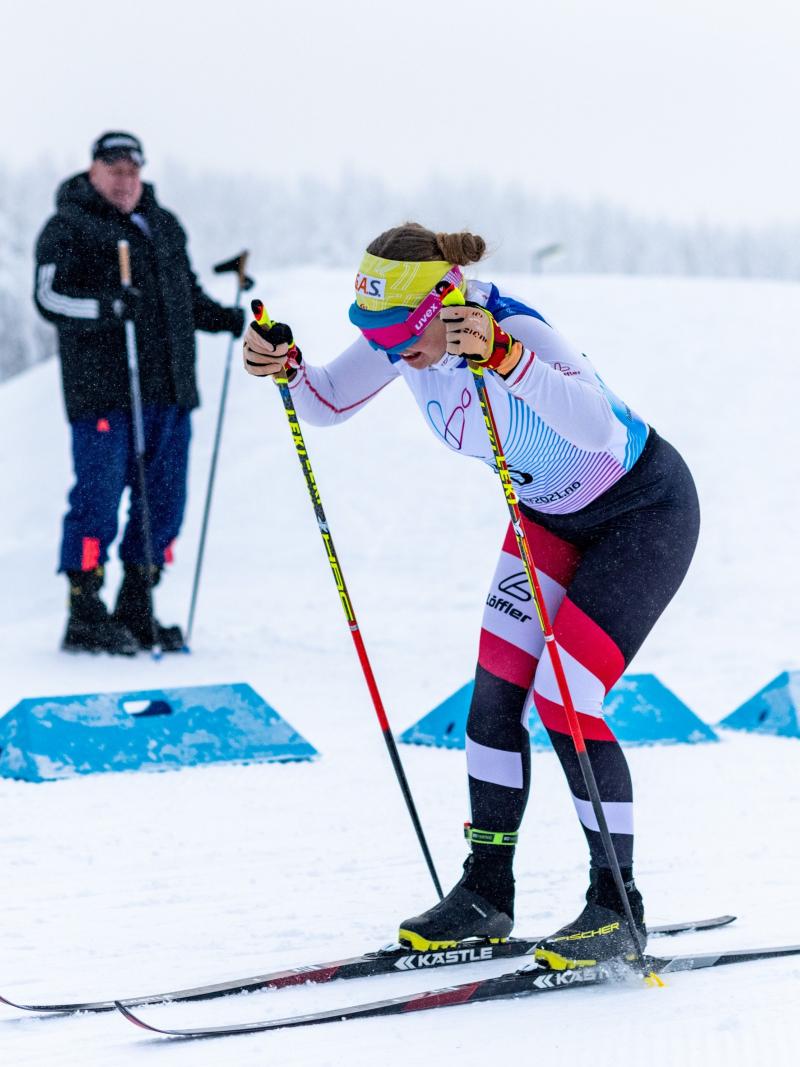 A female cross-country skier competing in the snow observed by a man in the background