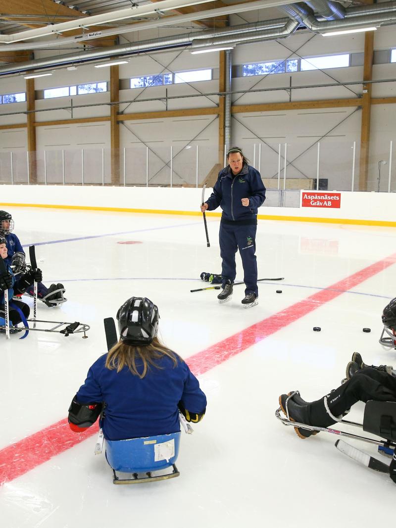A woman on an ice rink talking to a group of seven people on Para ice hockey sledges 
