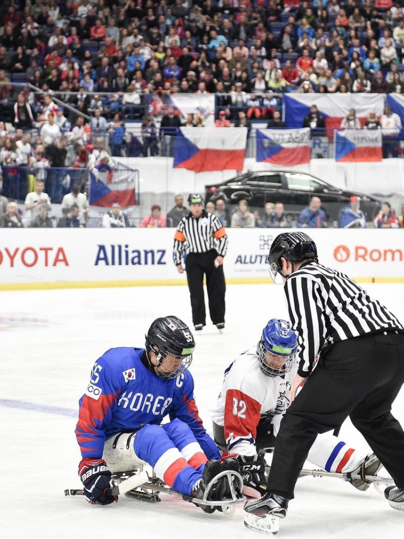 A referee throwing the puck in front of two players to start a Para ice hockey game in a crowded arena