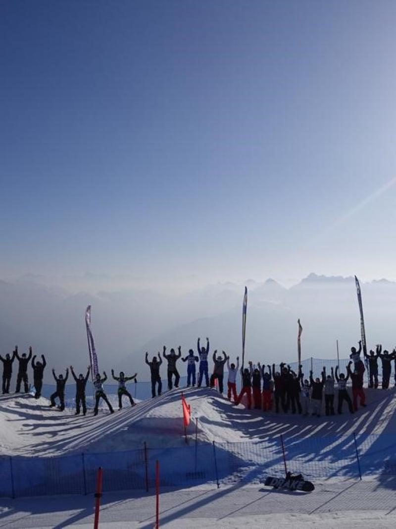 Snowboarders line up along a border-cross slope with their arms raised