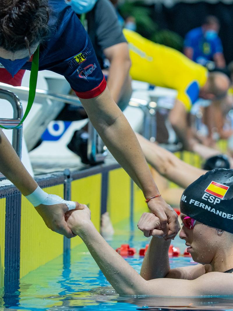 A woman outside a swimming pool grabbing the arms of a female Para swimmer inside the water