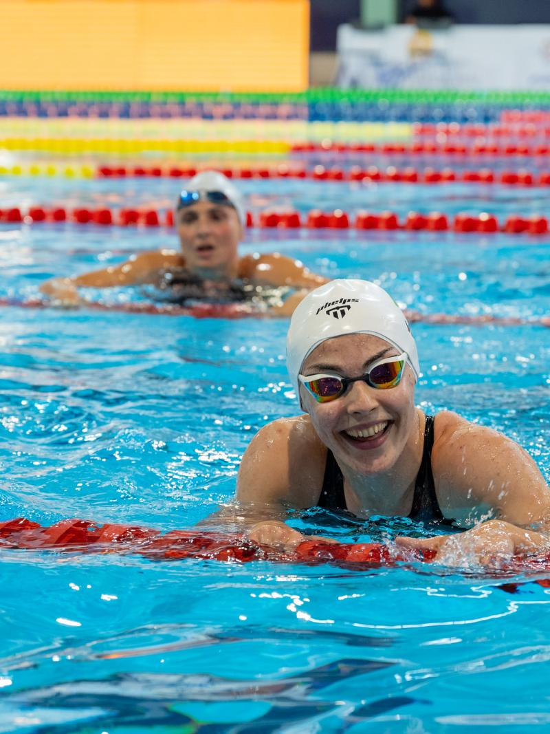 A young female swimmer with a white cap smiling in a swimming pool with another woman in the background