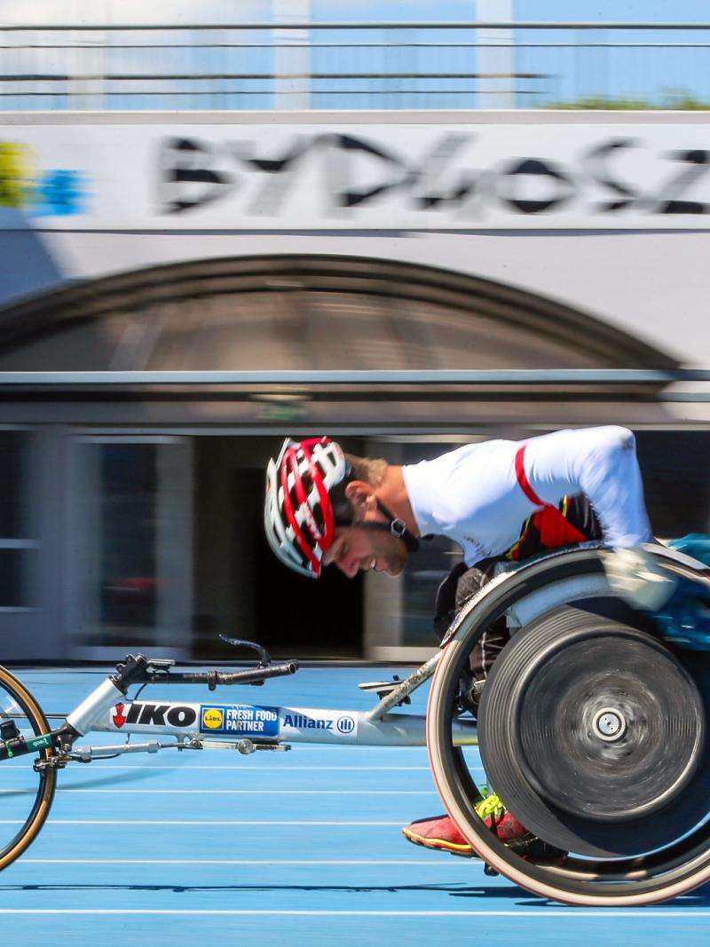 A man in a racing wheelchair on a blue athletics track passing in front of a gate